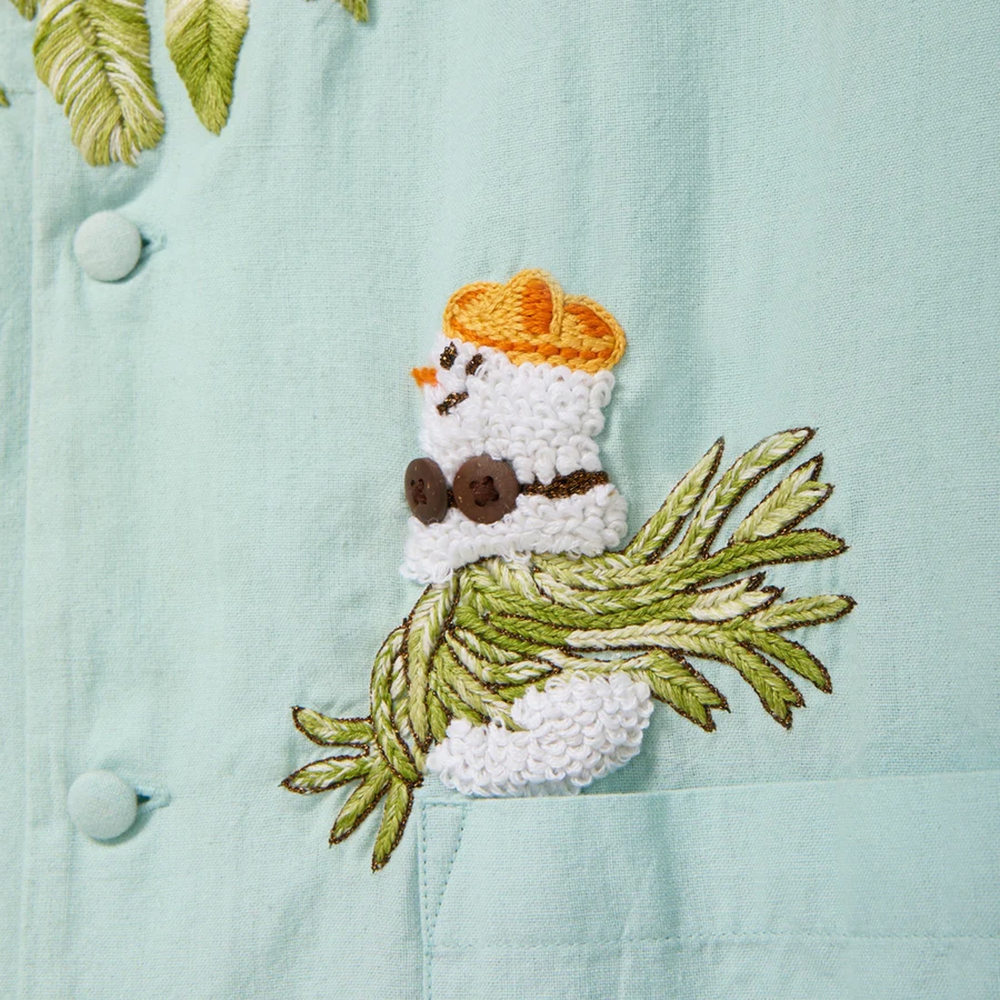 Close-up of a light blue cabana shirt showing embroidered artwork of a small snowman wearing sunglasses and sitting on embroidered palm fronds above a front pocket.