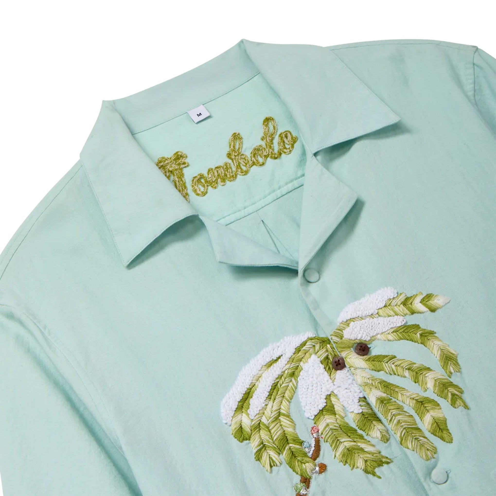 Close-up of a light blue cabana shirt showing the chain-stitched “Tombolo” logo at the collar and embroidered snowy palm tree detailing on the front.