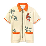 Short-sleeve zip-up shirt in cream terry cloth with bold orange accents at the collar, sleeve cuffs, and pocket openings. The shirt features embroidered green ivy vines and a red tiger in mid-pounce on the chest. A metal zipper with a circular pull runs down the center.