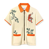 Short-sleeve zip-up shirt in cream terry cloth with bold orange accents at the collar, sleeve cuffs, and pocket openings. The shirt features embroidered green ivy vines and a red tiger in mid-pounce on the chest. A metal zipper with a circular pull runs down the center.