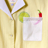 Close-up of a light yellow seersucker shirt with a white collar and pocket. The shirt features an embroidered lime slice and red chili pepper above the pocket, which is trimmed with beaded detailing.