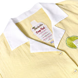 Close-up of light yellow textured shirt with a white collar and embroidered lime slice. The inner label is shaped like a cocktail shaker and reads 'Tombolo Crop Cut' with a playful margarita recipe printed below.