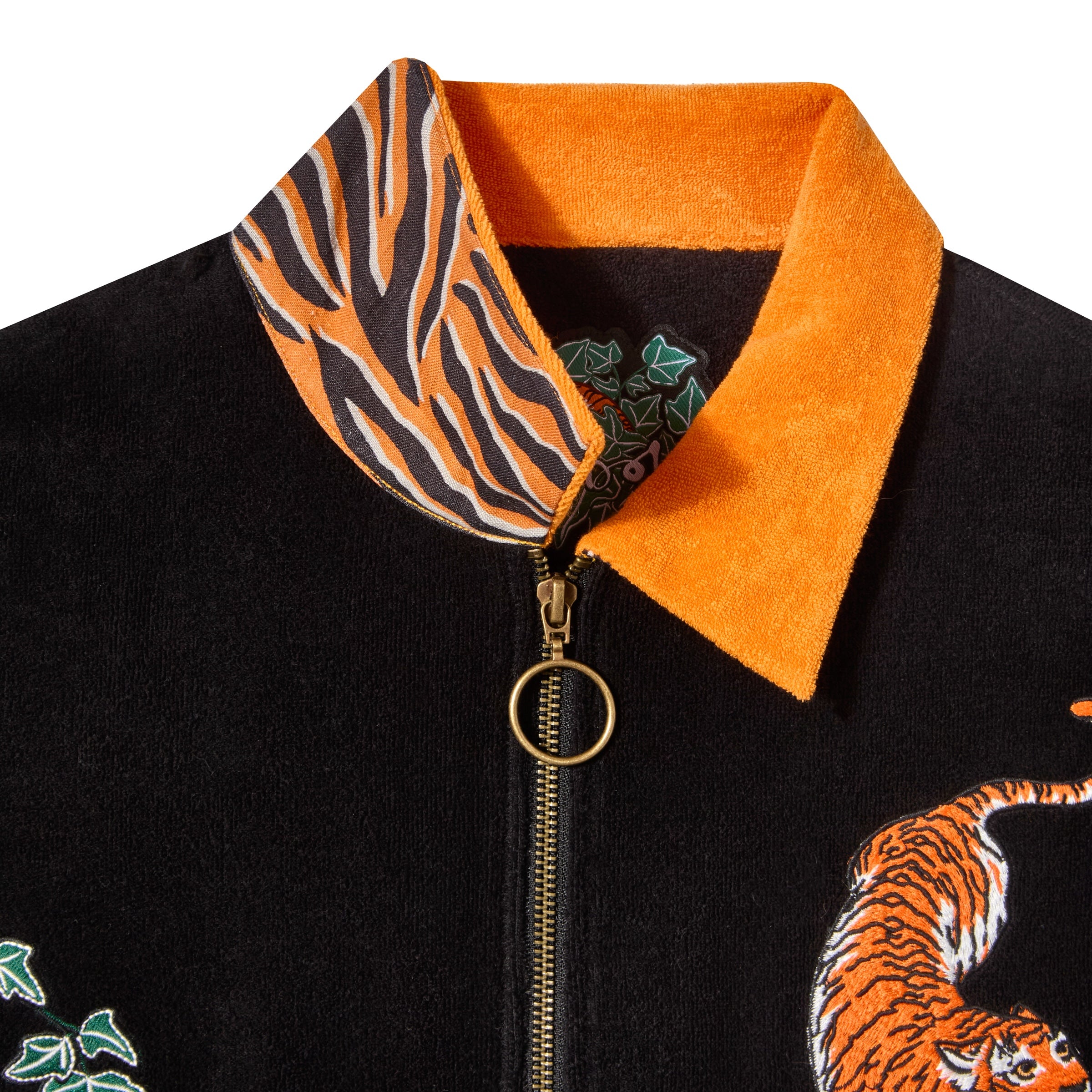 Close-up of a black terry cloth shirt with a bright orange collar. The inner collar features a tiger stripe pattern. Visible gold zipper with circular pull, partial tiger embroidery, and green ivy detailing on the shirt.