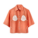 Orange short-sleeve button-up shirt with a relaxed fit. The shirt features an embroidered design resembling a sand dollar bikini top, with two large cream-colored sand dollars at the chest and shell bead strands extending from the shoulders, across the chest, and down the sides.