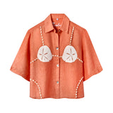 Orange short-sleeve button-up shirt with a relaxed fit. The shirt features an embroidered design resembling a sand dollar bikini top, with two large cream-colored sand dollars at the chest and shell bead strands extending from the shoulders, across the chest, and down the sides.