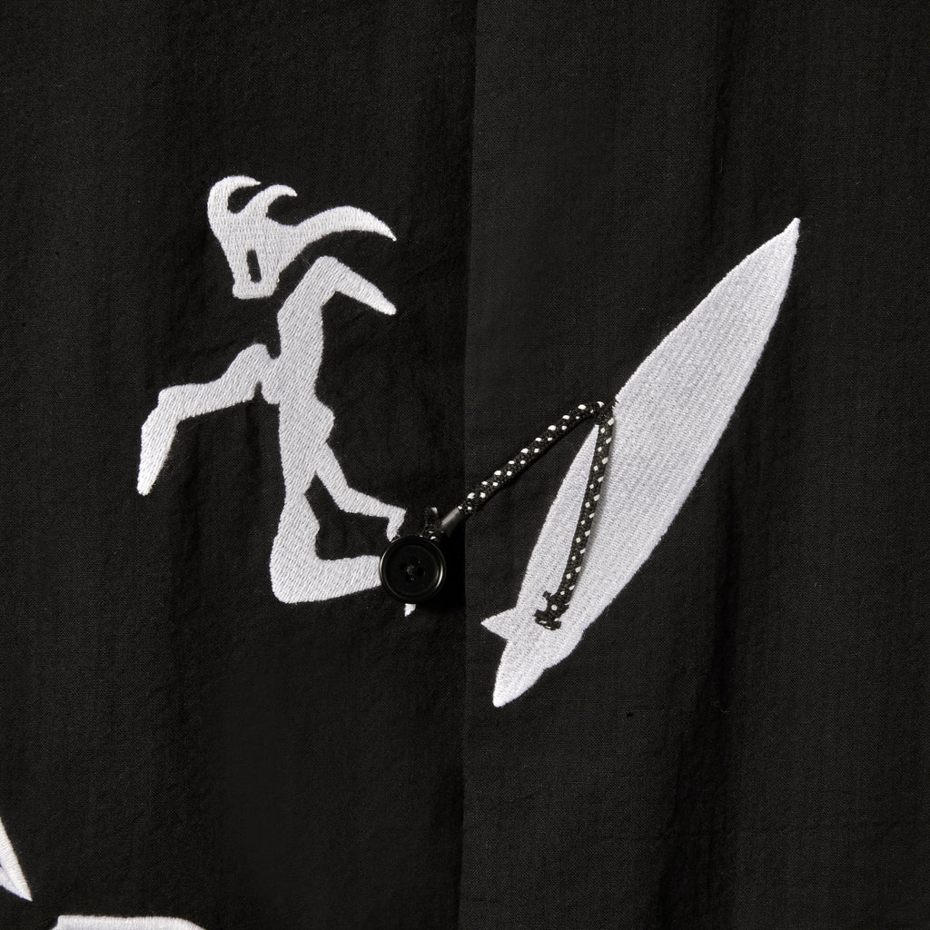 Close-up image of the embroidered surfer, attached to his board with an ankle leash. The ankle leash appears as a small loop to button the shirt.