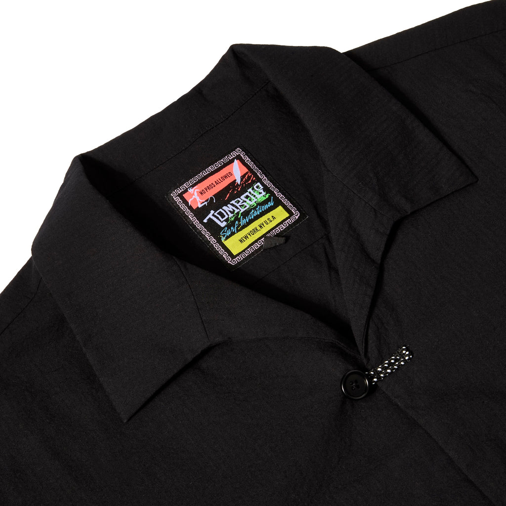 Close-up of a black shirt collar with a colorful Tombolo label reading 'Surf Invitational' and a black button with a braided loop closure below it.