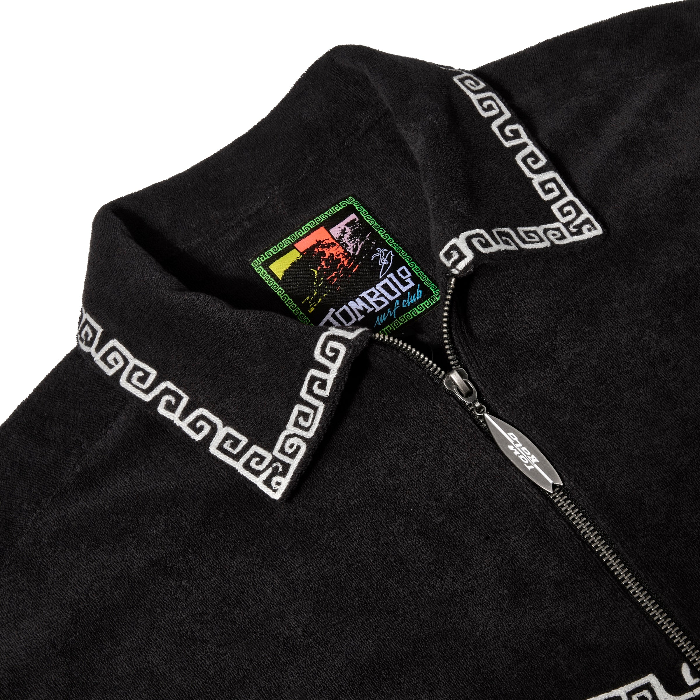 Close-up of black shirt collar with white trim, a Tombolo Surf Club label inside, and a silver surfboard-shaped zipper pull.