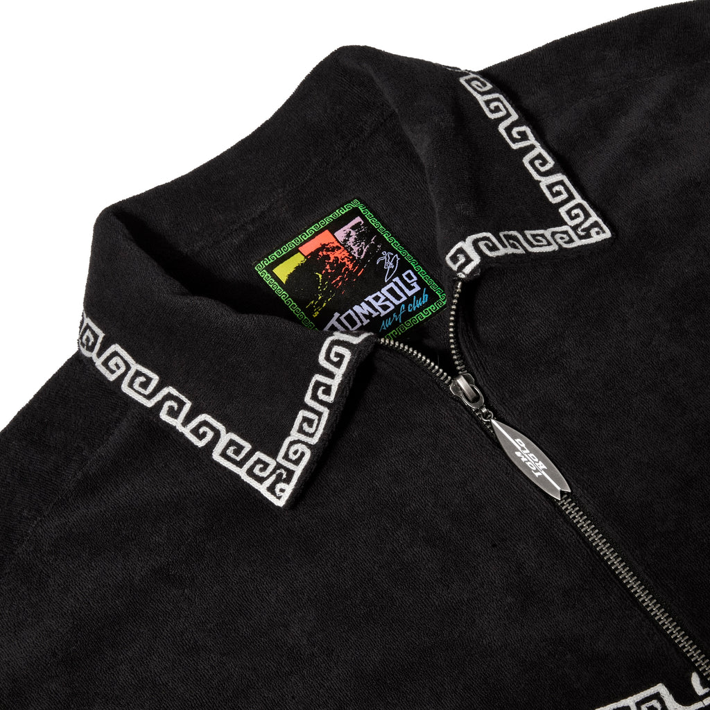 Close-up of black shirt collar with white trim, a Tombolo Surf Club label inside, and a silver surfboard-shaped zipper pull.