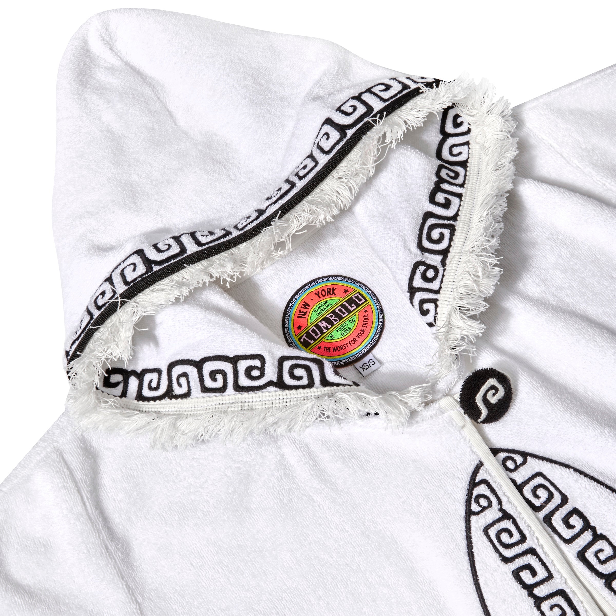 Close-up of white terry cloth hooded beach towel top showing frayed edges, black embroidered trim, and a colorful Tombolo brand label inside the collar.
