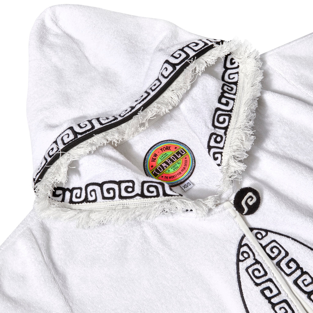 Close-up of white terry cloth hooded beach towel top showing frayed edges, black embroidered trim, and a colorful Tombolo brand label inside the collar.