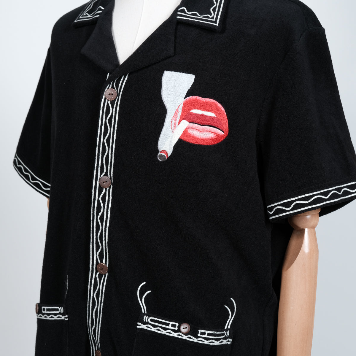 Close-up of a black short-sleeve shirt on a mannequin, featuring cream embroidered trim and a bold red lips and cigarette graphic on the chest. Brown buttons and detailed pocket embroidery complete the design.