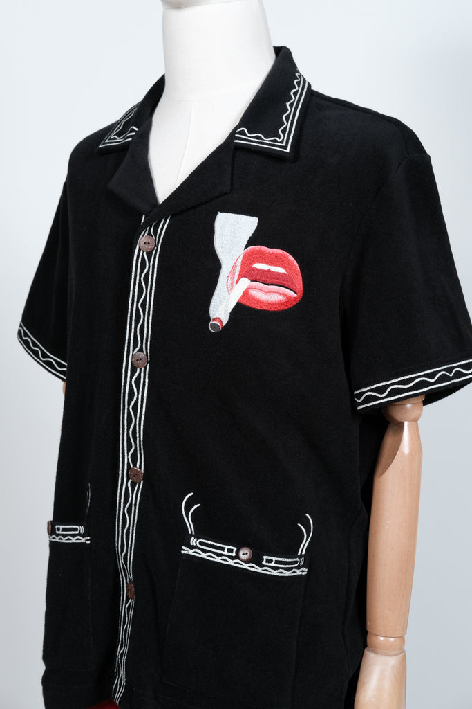 Close-up of a black short-sleeve shirt on a mannequin, featuring cream embroidered trim and a bold red lips and cigarette graphic on the chest. Brown buttons and detailed pocket embroidery complete the design.
