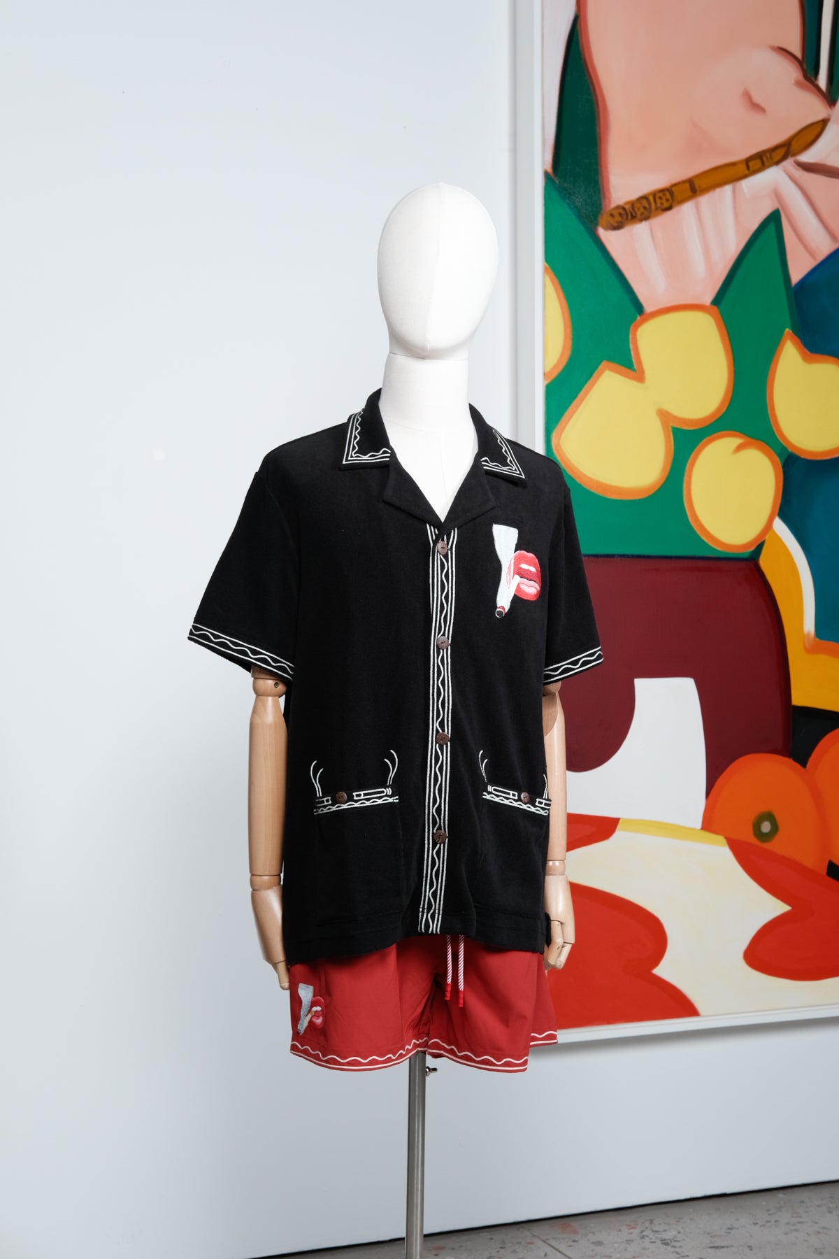 Mannequin dressed in a black embroidered cabana shirt and red swim trunks, both featuring a matching lips and cigarette motif. 