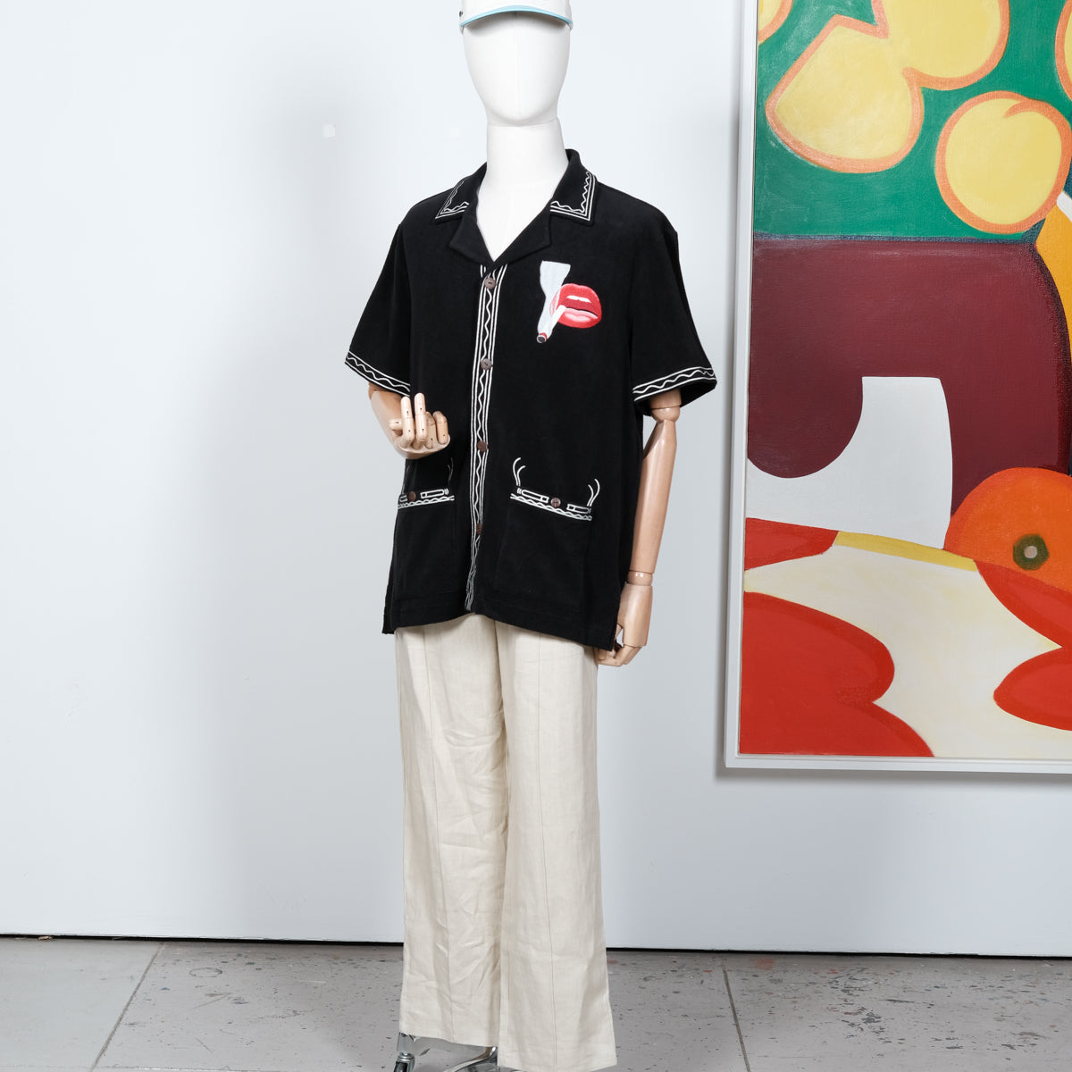 Black short-sleeve shirt displayed on a mannequin, featuring cream embroidered trim and a red lips and cigarette graphic on the left chest. Styled with off-white pants and a matching embroidered cap.