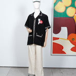 Black short-sleeve shirt displayed on a mannequin, featuring cream embroidered trim and a red lips and cigarette graphic on the left chest. Styled with off-white pants and a matching embroidered cap.