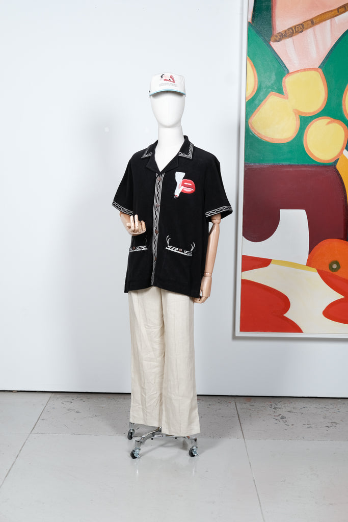 Black short-sleeve shirt displayed on a mannequin, featuring cream embroidered trim and a red lips and cigarette graphic on the left chest. Styled with off-white pants and a matching embroidered cap.