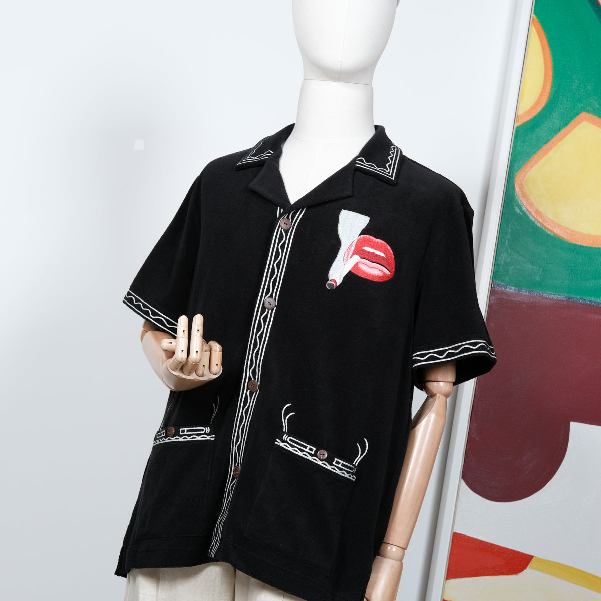 Black short-sleeve shirt displayed on a mannequin, featuring cream embroidered trim and a red lips and cigarette graphic on the left chest. Styled with off-white pants and a matching embroidered cap.
