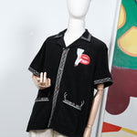 Black short-sleeve shirt displayed on a mannequin, featuring cream embroidered trim and a red lips and cigarette graphic on the left chest. Styled with off-white pants and a matching embroidered cap.