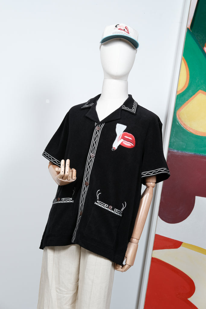 Black short-sleeve shirt displayed on a mannequin, featuring cream embroidered trim and a red lips and cigarette graphic on the left chest. Styled with off-white pants and a matching embroidered cap.