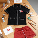 Black short-sleeve shirt and red swim trunks laid flat on a wooden artist's table, surrounded by paintbrushes, tubes of paint, and printed artwork. Both garments feature matching embroidery of red lips with a lit cigarette.