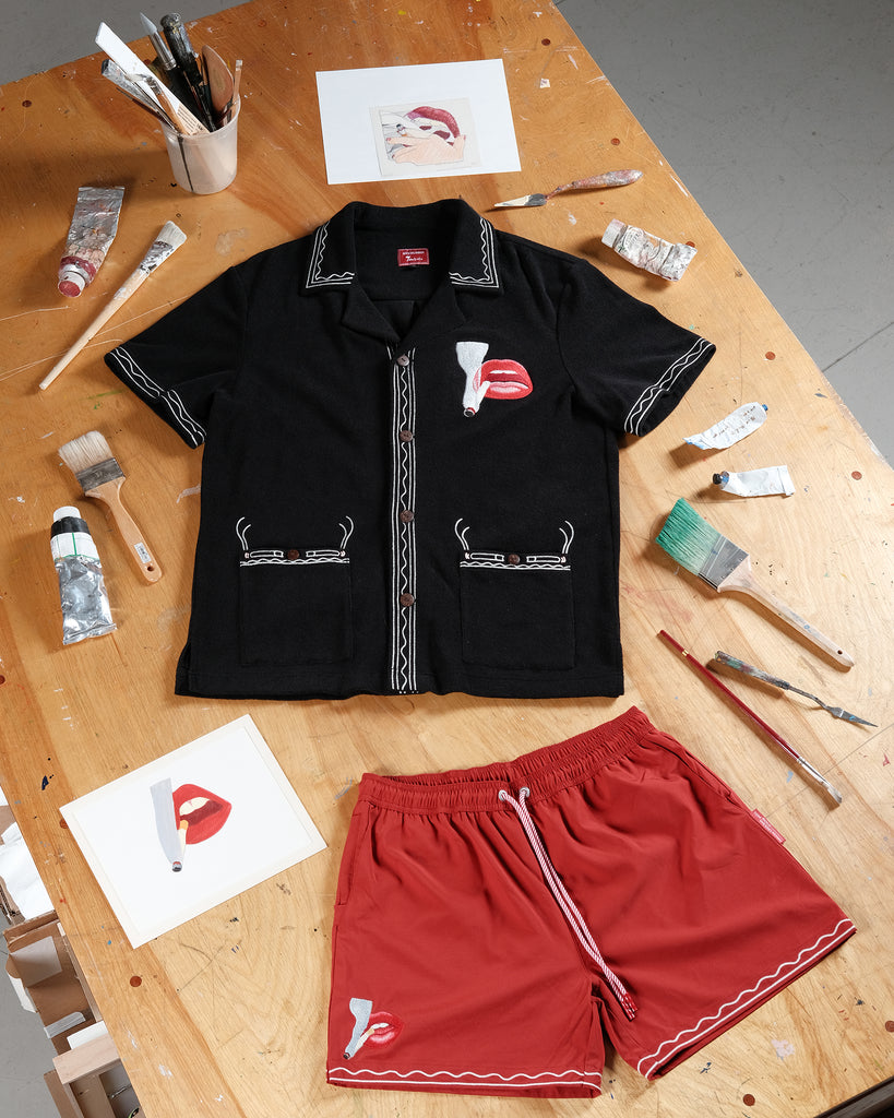 Black short-sleeve shirt and red swim trunks laid flat on a wooden artist's table, surrounded by paintbrushes, tubes of paint, and printed artwork. Both garments feature matching embroidery of red lips with a lit cigarette.