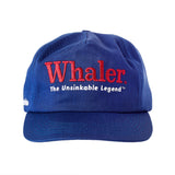 Blue five-panel cap with red embroidered text reading 'Whaler' and white text below reading 'The Unsinkable Legend.' Small white 'Tombolo' logo on the side of the hat.