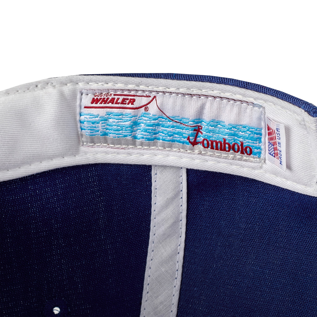 Close-up of the inside label on a blue five-panel cap, featuring a Boston Whaler boat graphic above blue waves and red 'Tombolo' text. A 'Made in USA' tag is visible beside it.