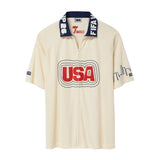 Cream short-sleeve terry cloth zip-front shirt with large red “USA” appliqué across the chest, navy contrast collar with white lettering, and embroidered skyline graphics on the sleeves.