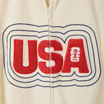 Close-up of cream terry cloth zip shirt showing large red “USA” appliqué across the chest with navy stitched outline and small FIFA trophy logo inside the letter A.
