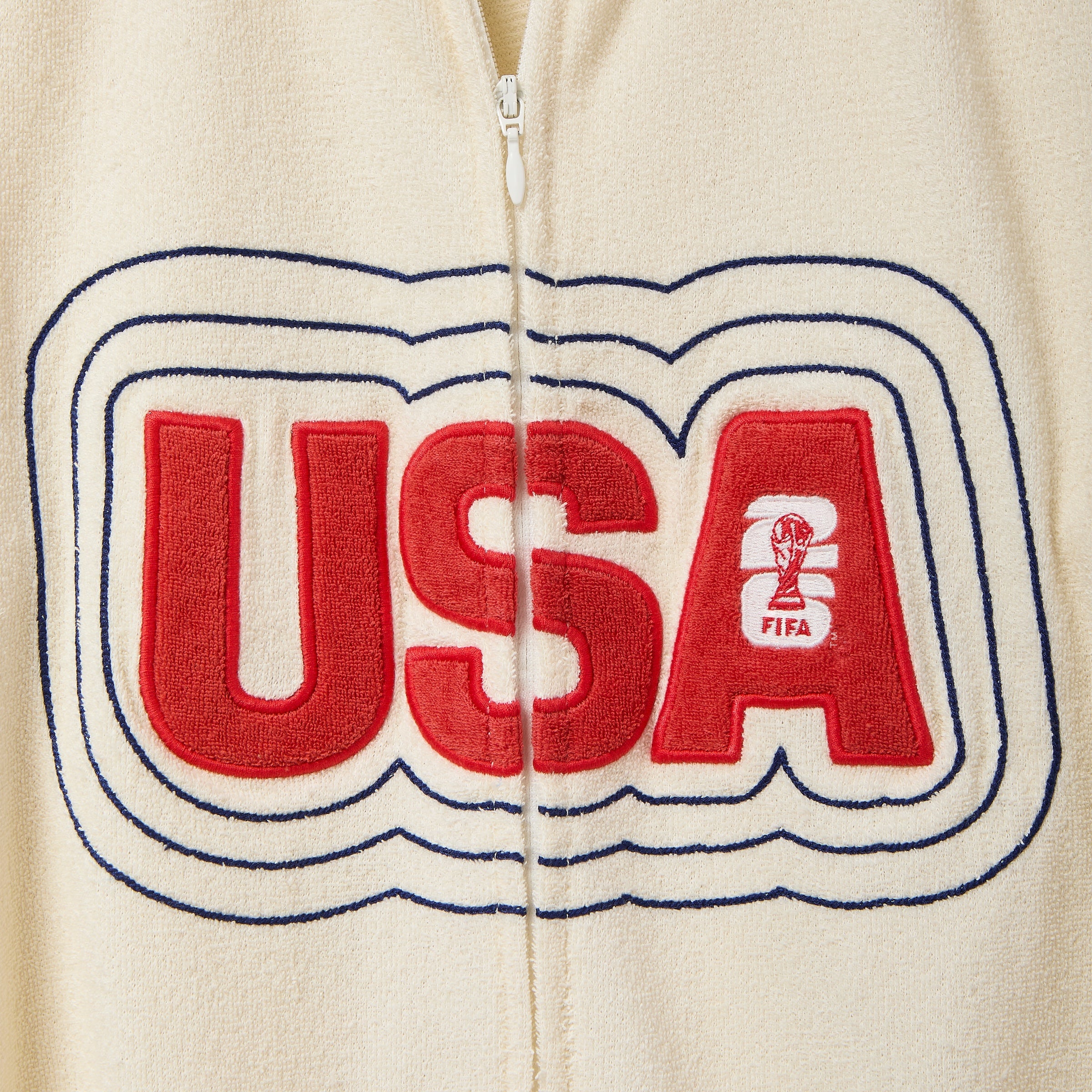 Close-up of cream terry cloth zip shirt showing large red “USA” appliqué across the chest with navy stitched outline and small FIFA trophy logo inside the letter A.