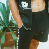 Close-up of a model wearing the green terry cloth shirt and matching shorts. The shirt has an embroidered pocket with two playing cards, and the shorts feature a vertical side stripe patterned with red and black playing card suits. His hand rests in his pocket.