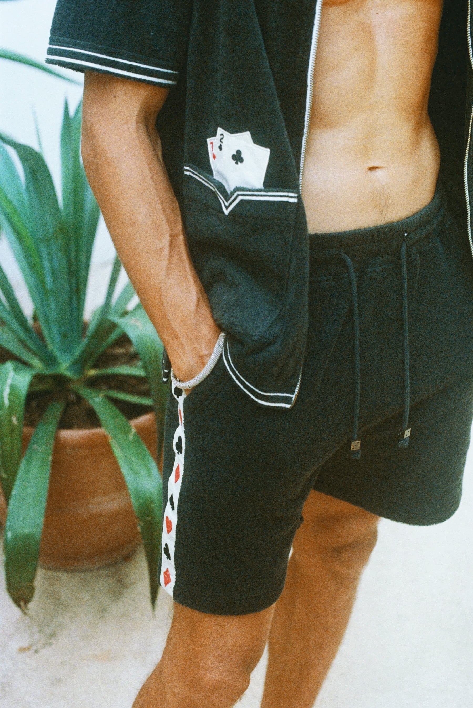 Close-up of a model wearing the green terry cloth shirt and matching shorts. The shirt has an embroidered pocket with two playing cards, and the shorts feature a vertical side stripe patterned with red and black playing card suits. His hand rests in his pocket.