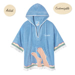 Sky blue terrycloth poncho with white fringe and trim, featuring an embroidered design of legs with red-painted toenails and waves along the hem. 'Tombolo' is stitched in white on the chest.