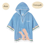 Sky blue terrycloth poncho with white fringe and trim, featuring an embroidered design of legs with red-painted toenails and waves along the hem. 'Tombolo' is stitched in white on the chest.