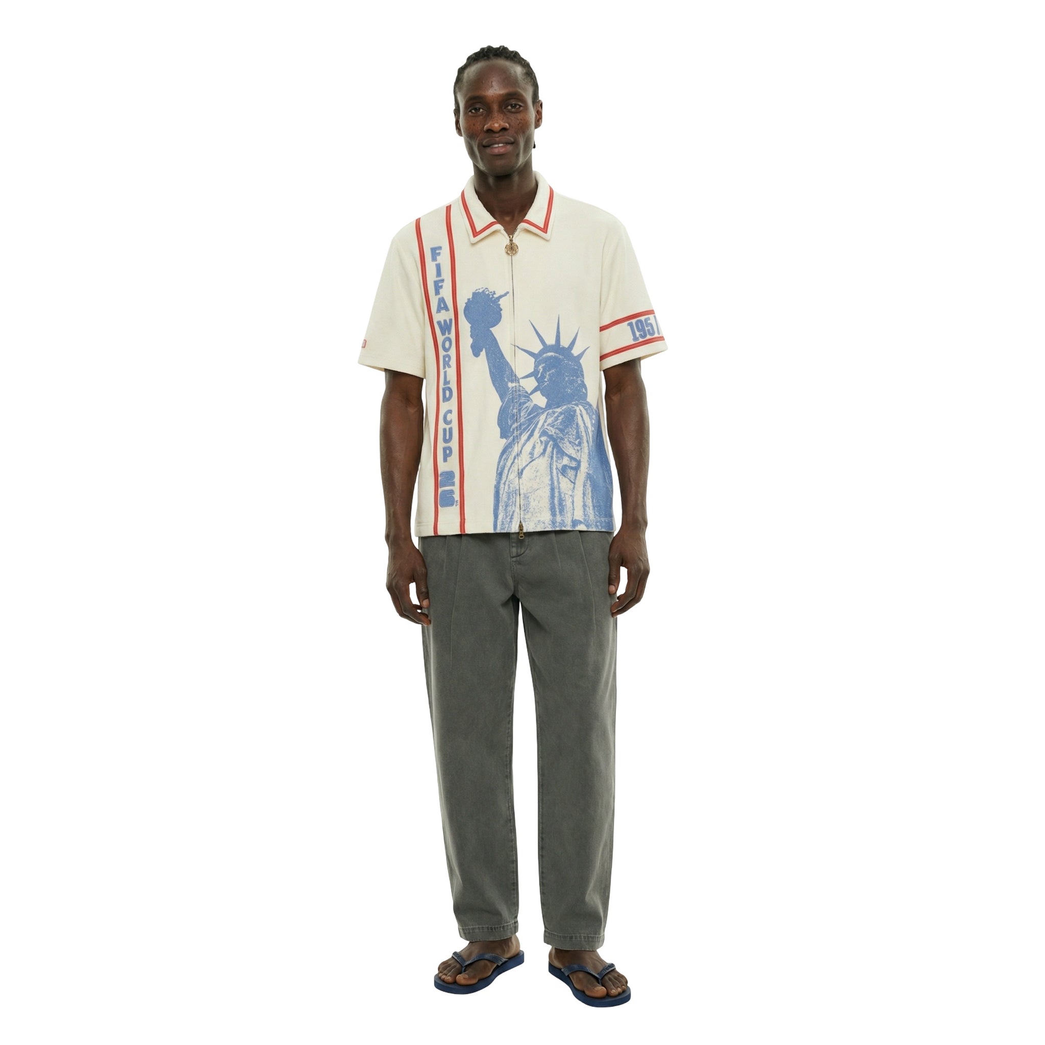 Model wearing a cream short-sleeve zip shirt with large blue Statue of Liberty graphic on the front, vertical FIFA World Cup lettering, and red contrast trim at the collar and sleeves.