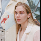Shot of a male model's 'Hermit Crabana' Cabana and its appliquéd and edge-embroidered hermit crab shaped into the placket, in addition to a female model with long blonde hair and a 'conched out' cabana beside him, shot on a background of blue skies and palm fronds.