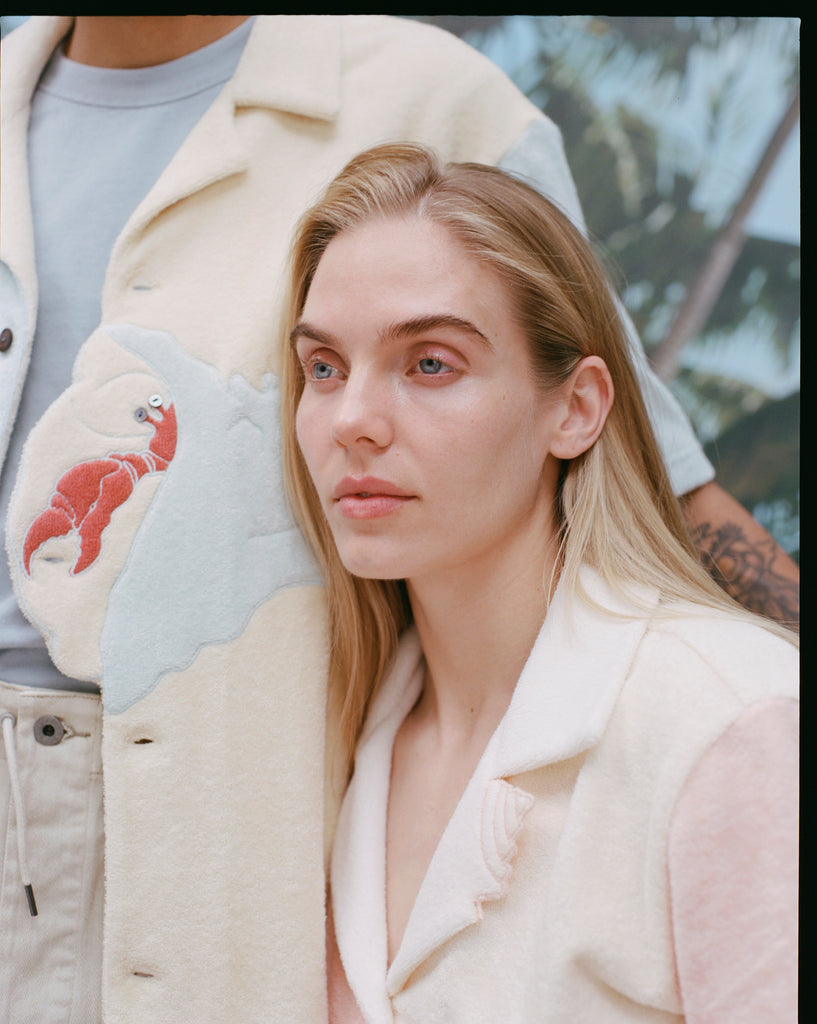 Shot of a male model's 'Hermit Crabana' Cabana and its appliquéd and edge-embroidered hermit crab shaped into the placket, in addition to a female model with long blonde hair and a 'conched out' cabana beside him, shot on a background of blue skies and palm fronds.