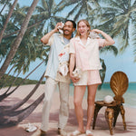 Male model wearing the 'Hermit Crabana' Cabana with khakis and beige slippers while holding a conch to his ear alongside a female model wearing the 'conched out' cabana with matching 'conched out' shorts, shot on a background with a beach, palm trees, and blue skies. 