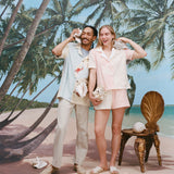 Male model wearing the 'Hermit Crabana' Cabana with khakis and beige slippers while holding a conch to his ear alongside a female model wearing the 'conched out' cabana with matching 'conched out' shorts, shot on a background with a beach, palm trees, and blue skies. 