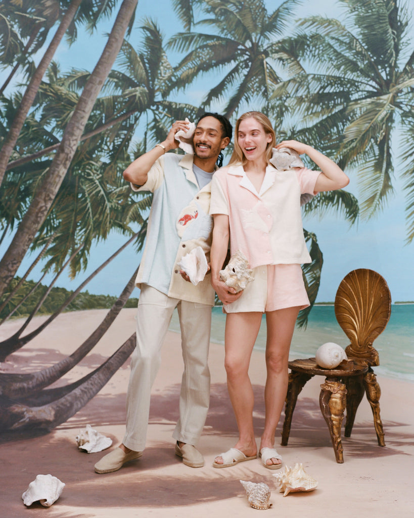 Male model wearing the 'Hermit Crabana' Cabana with khakis and beige slippers while holding a conch to his ear alongside a female model wearing the 'conched out' cabana with matching 'conched out' shorts, shot on a background with a beach, palm trees, and blue skies. 