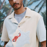 Male model wearing the 'Hermit Crabana' Cabana with a light blue shirt underneath, shot on a background with palm fronds and blue skies.