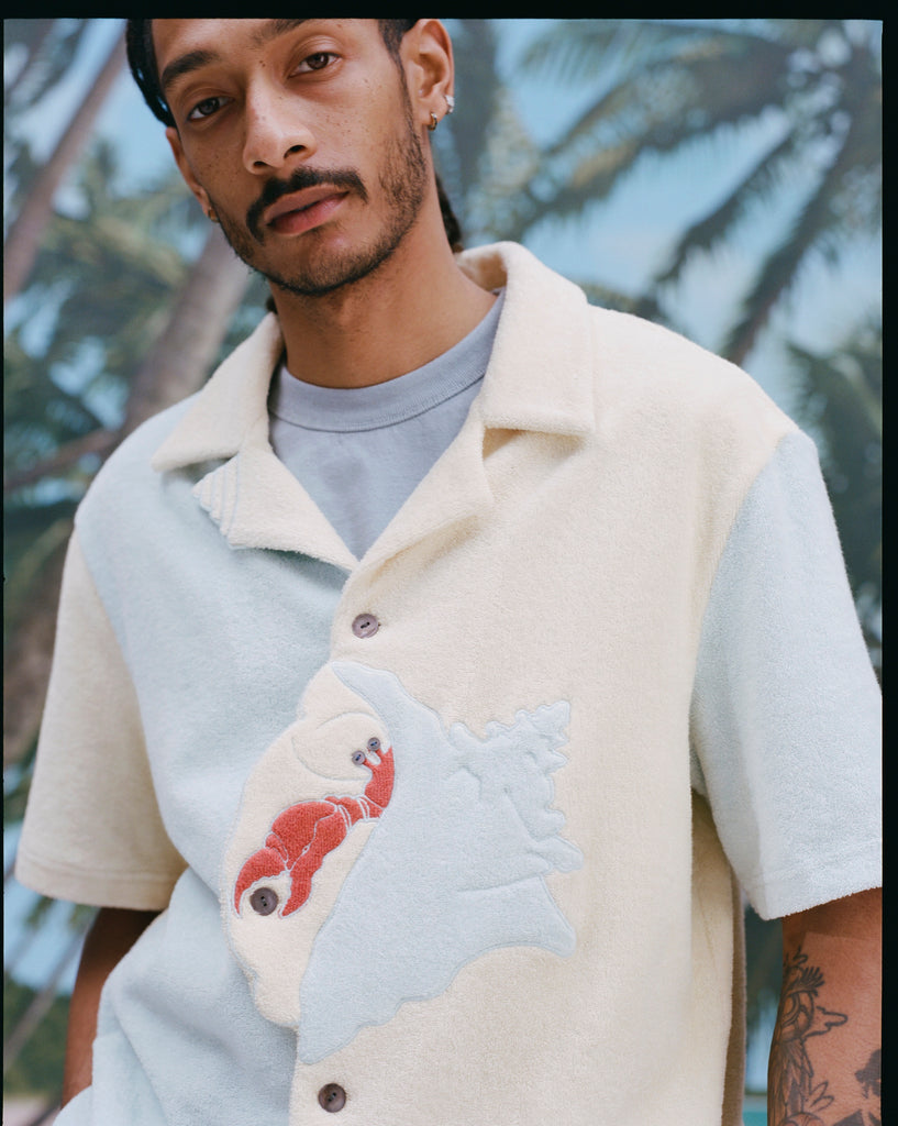 Male model wearing the 'Hermit Crabana' Cabana with a light blue shirt underneath, shot on a background with palm fronds and blue skies.