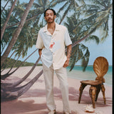 Male model with dreadlocks wearing the 'Hermit Crabana' Cabana with khaki pants and beige slippers, shot on a background of palm trees, blue skies, and a beach.