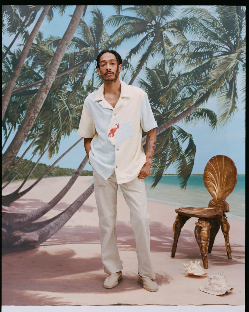 Male model with dreadlocks wearing the 'Hermit Crabana' Cabana with khaki pants and beige slippers, shot on a background of palm trees, blue skies, and a beach.