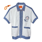 Short-sleeved zip-up terry cloth shirt in white with blue trim and piping. Features two front pockets, a ring zipper pull, and a chest patch with “The Explorers Club NYC,” the letters “E” and “C,” and a small shark illustration. An orange whistle on a coiled wristband is shown beside the shirt.