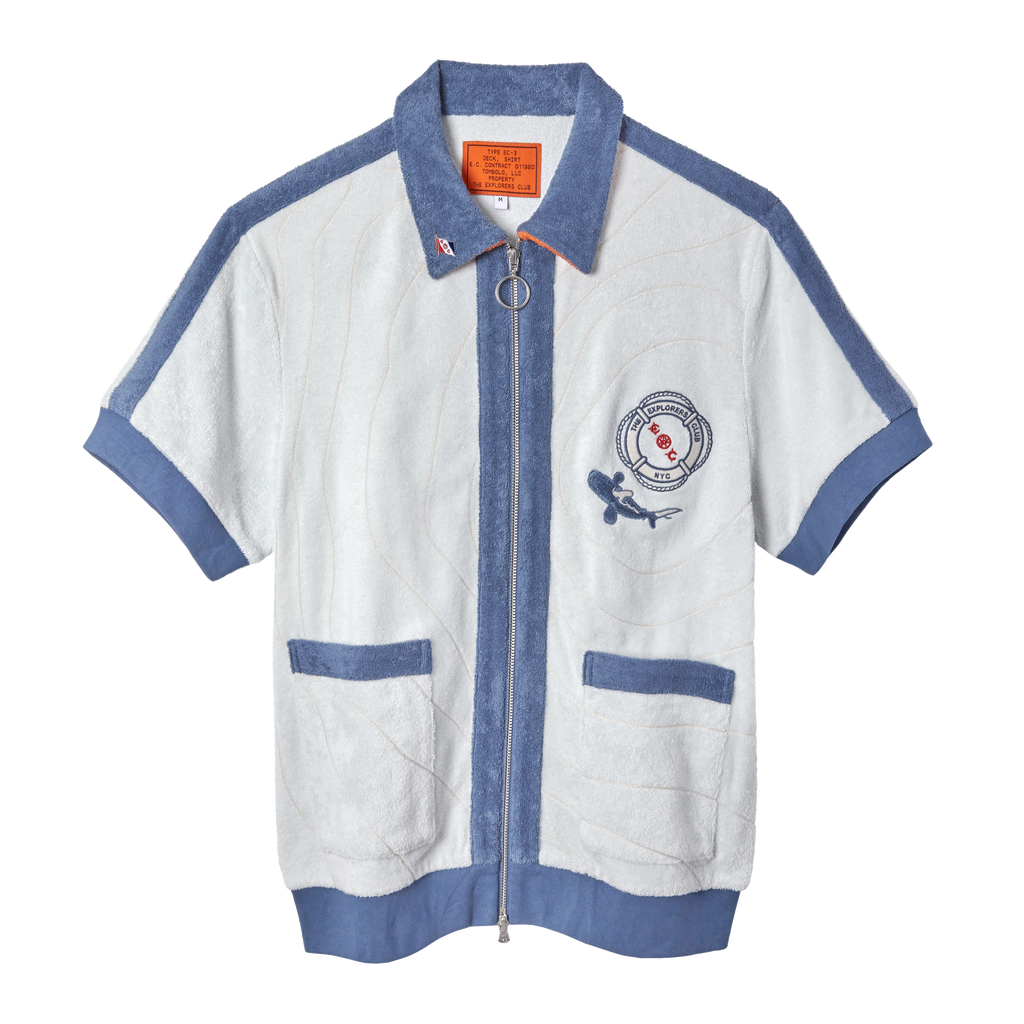 Short-sleeved zip-up terry cloth shirt in white with blue trim and piping. Features two front pockets, a ring zipper pull, and a chest patch with “The Explorers Club NYC,” the letters “E” and “C,” and a small shark illustration.