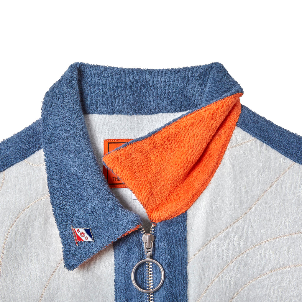 Close-up of a terry cloth shirt collar in blue with a bright orange underside. The shirt features a metal zipper with a round pull ring and a small embroidered flag patch with the letters “E” and “C” on the collar.