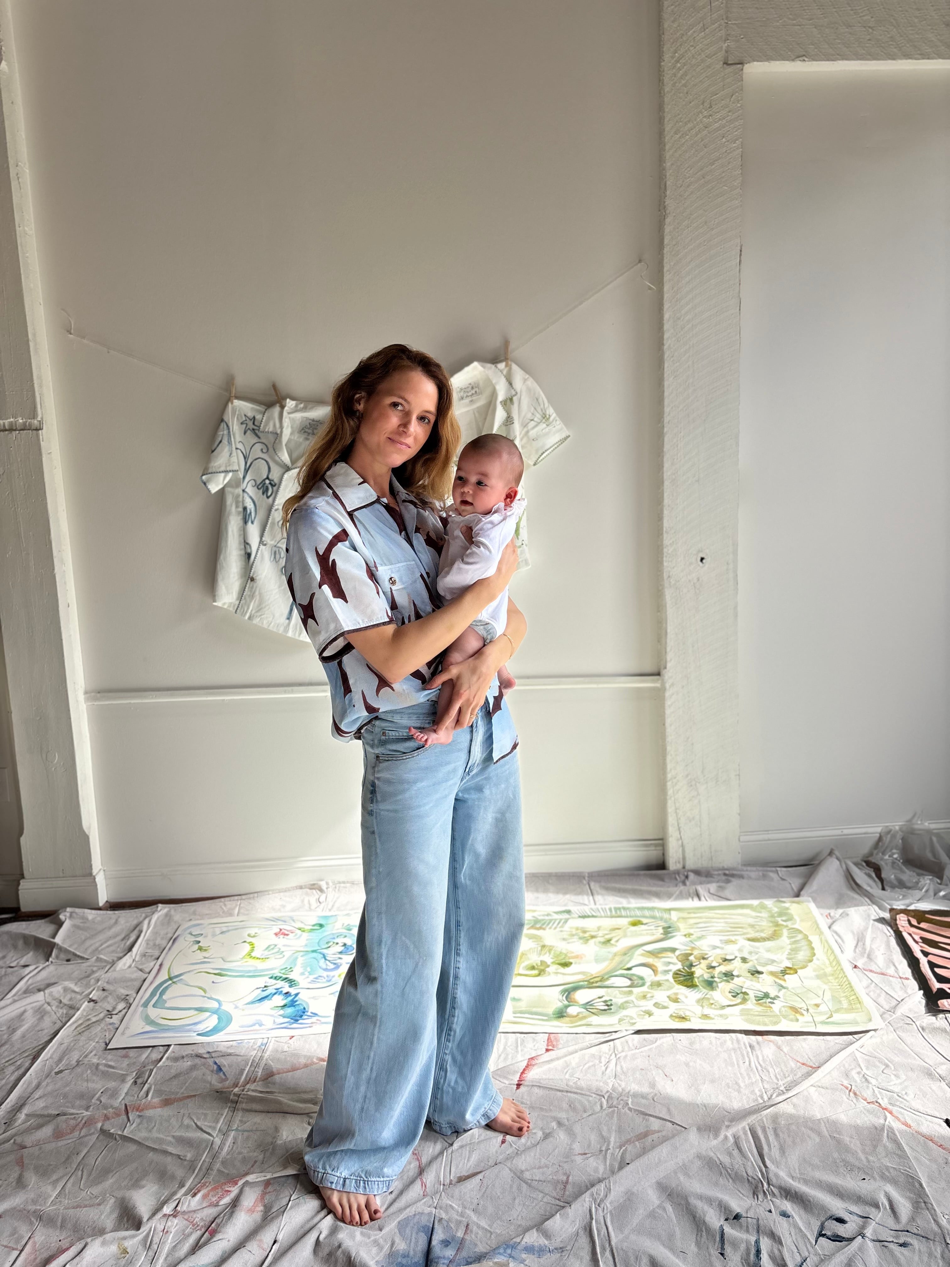 Mary Ball standing in her studio with her baby in her arms and smiling at the camera. 