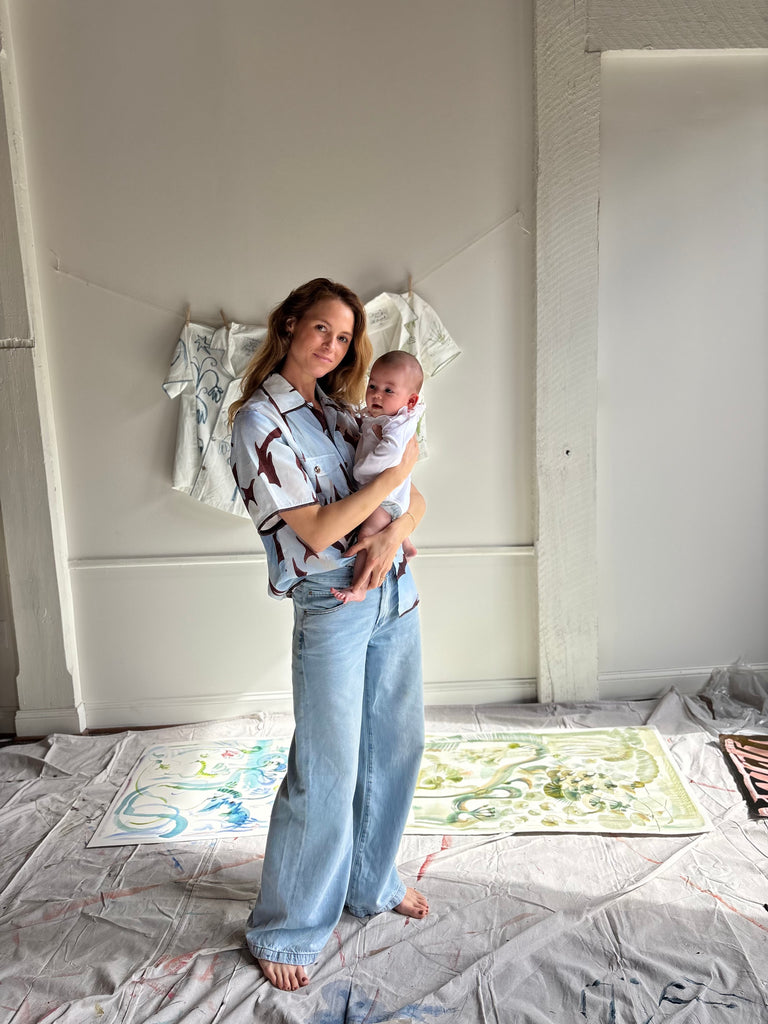Mary Ball standing in her studio with her baby in her arms and smiling at the camera. 